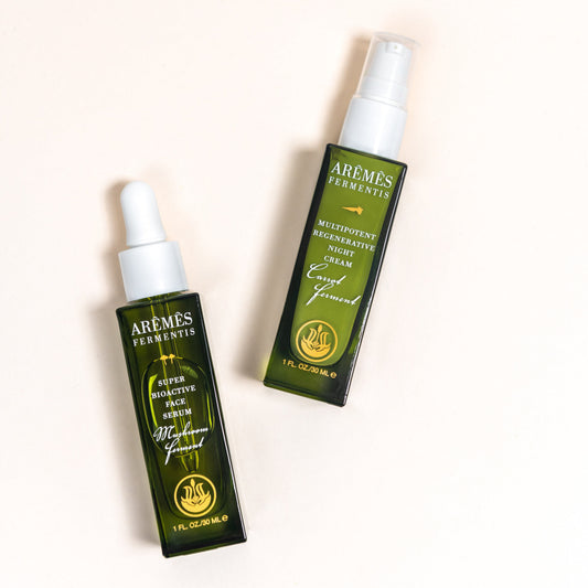 Two Aremés Ferments skincare bottles on a light cream background: a dark green dropper bottle labeled 'Cica Soothing Serum' and a matching spray bottle labeled 'Moisturizing Restorative Serum'.